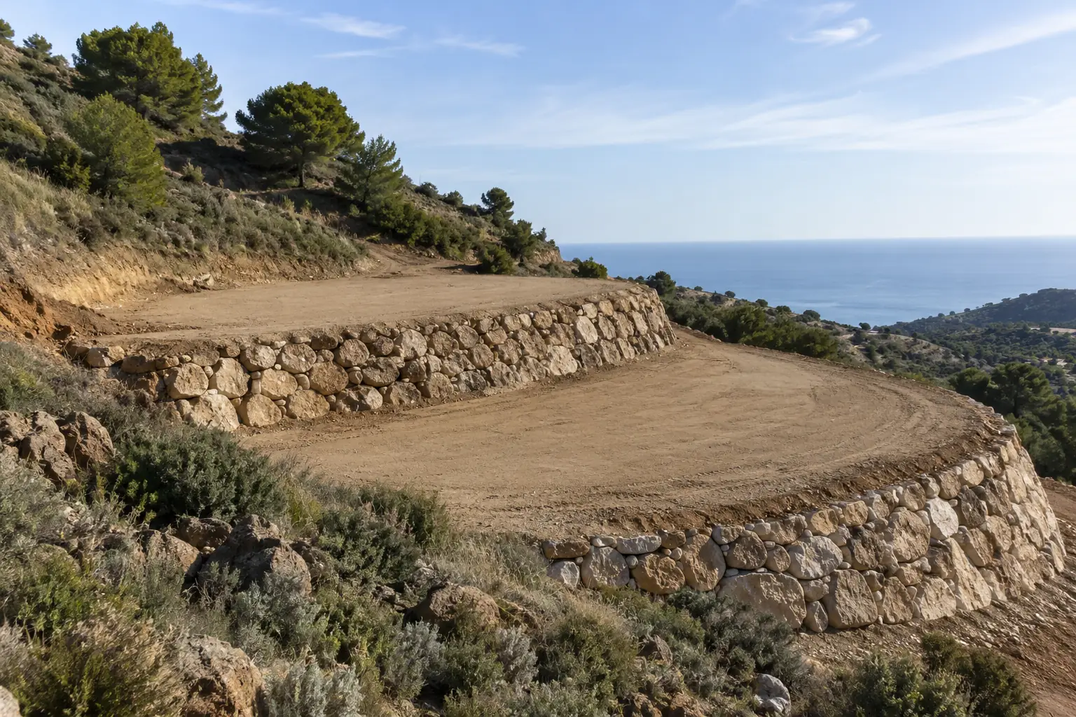 Ladera transformada en dos amplios bancales planos de tierra compactada, contenidos por muros de escollera, sin maquinaria, vegetación mediterránea.