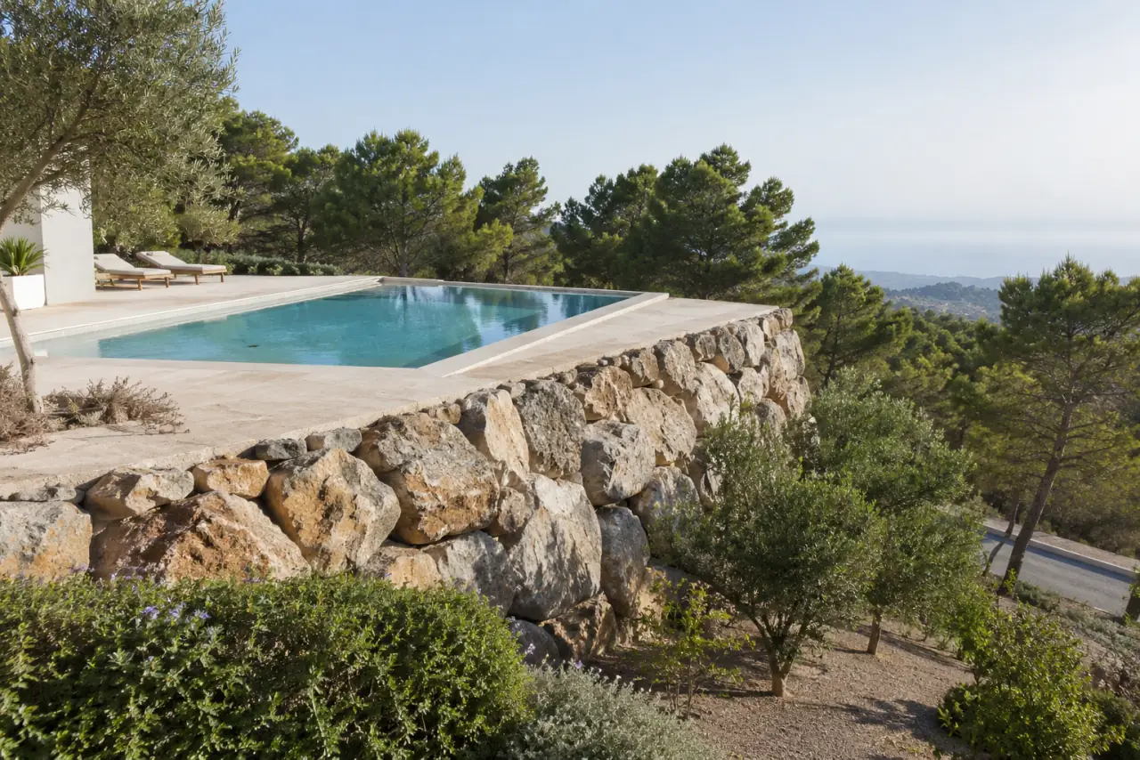 Piscina de obra sobre un bancal que sostiene una escollera de piedra natural, pavimento claro y pinos al fondo.