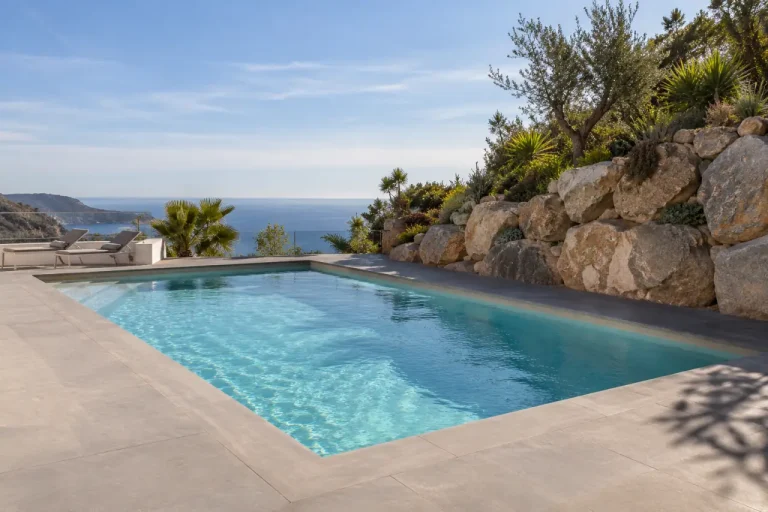 Piscina con muro de escollera — en Nerja Piscina de obra con pavimento claro y un muro de escollera al fondo.